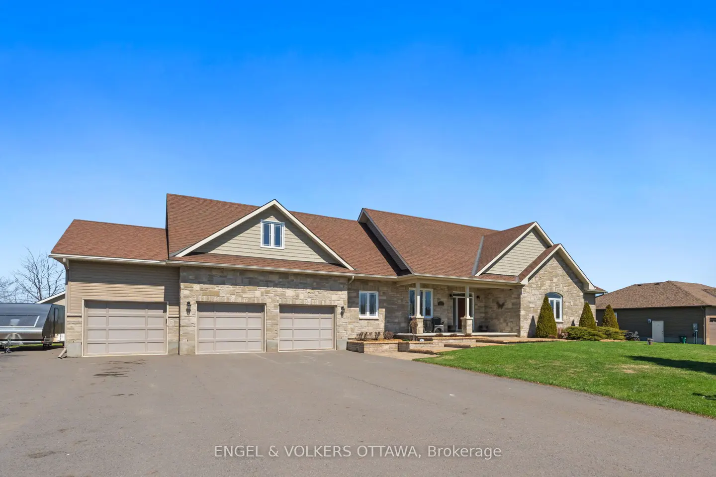 A large, tan stone house with a brown roof and three-car garage under a clear blue sky.