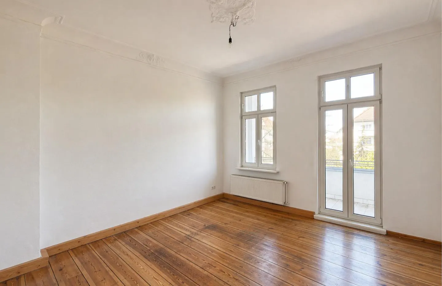 Empty room with white walls, wood floors, and trim. Two white windows let in natural light. A radiator sits beneath one window.