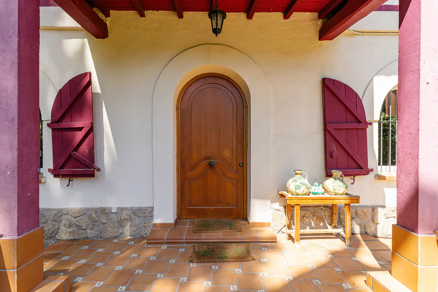 Exterior view of a home with a wooden arched door, red shutters, and a small table with vases.