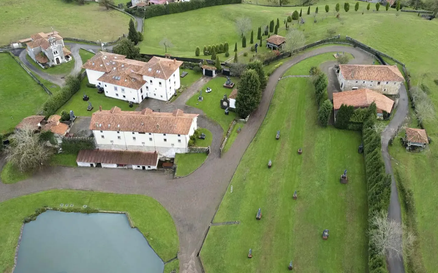 Aerial view of a large estate with white buildings, terracotta roofs, a pond, and sculptures scattered across green lawns.