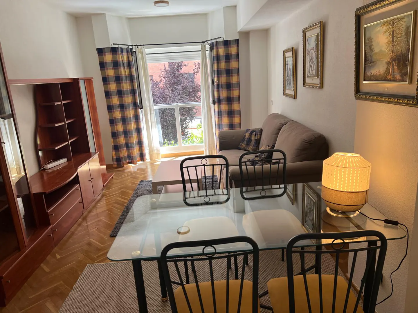 Living room with a glass table, black chairs, brown sofa, and wooden cabinet. Plaid curtains frame a bright window.