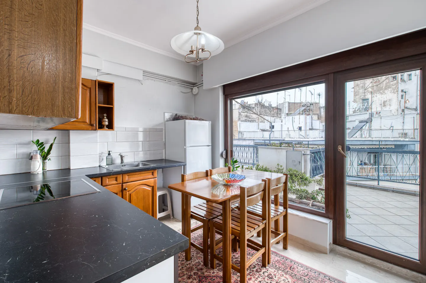 A bright kitchen with wood cabinets, black countertops, and a dining table. A large window overlooks a balcony with city views.
