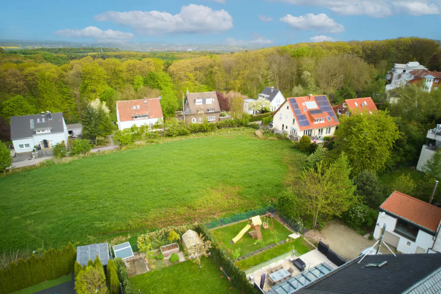 Aerial view of houses with red and gray roofs, surrounded by green trees and a large green field under a blue sky.