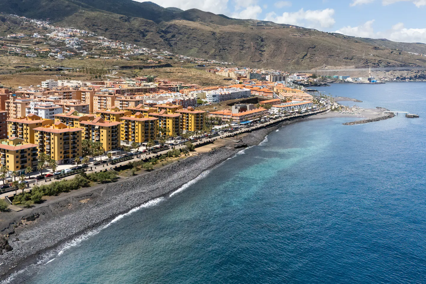Coastal view of yellow buildings lining a black sand beach with blue ocean and mountains in the background.