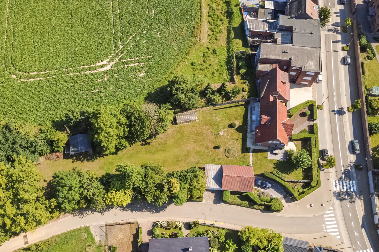 Aerial view of a red-roofed house with a large green lawn next to a green field and a road with cars.