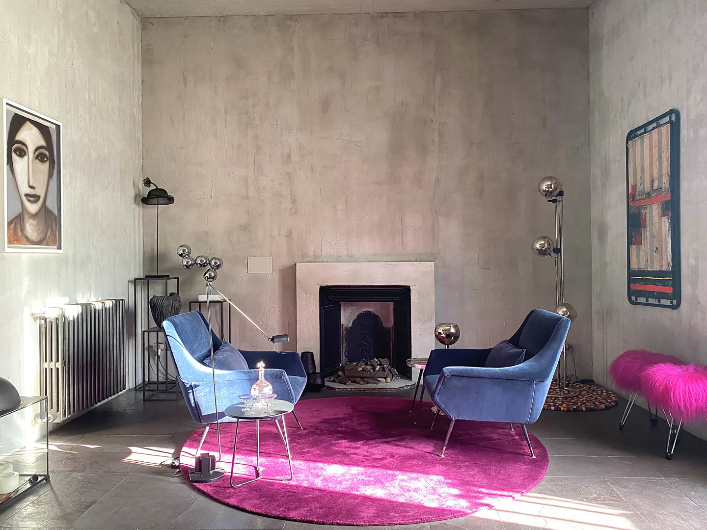 Living room with two blue chairs, a pink rug, and a fireplace. Art hangs on the gray walls.