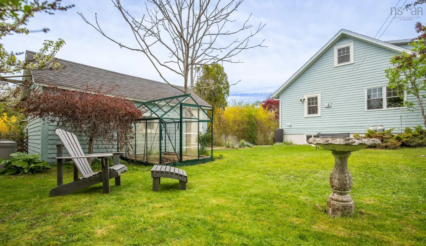 Backyard view with a green greenhouse, Adirondack chair, and birdbath on a green lawn. Two light blue houses are in the background.