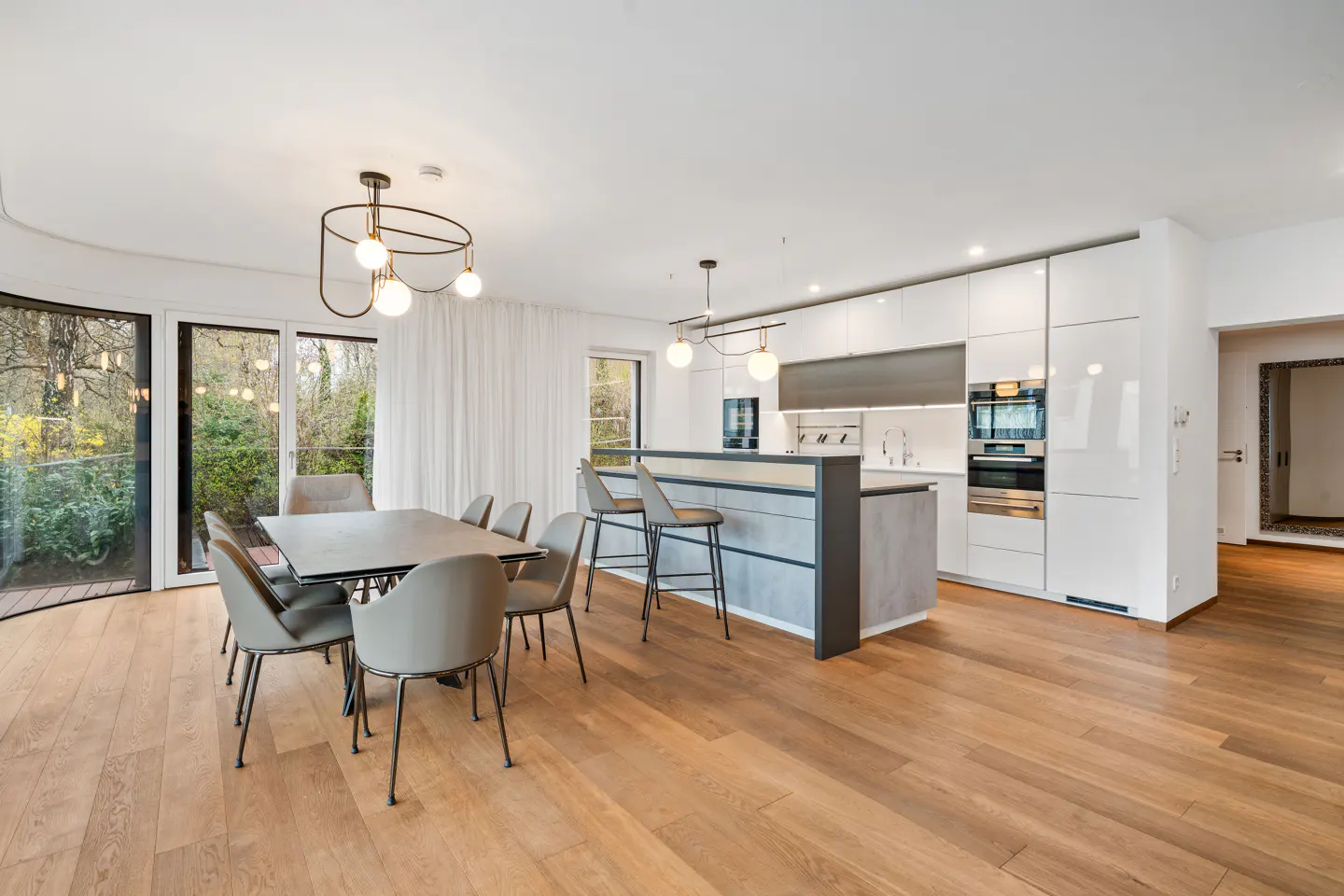 Bright, open-plan kitchen and dining area with wood floors. A table with six chairs sits near glass doors. A kitchen island and white cabinets are visible.