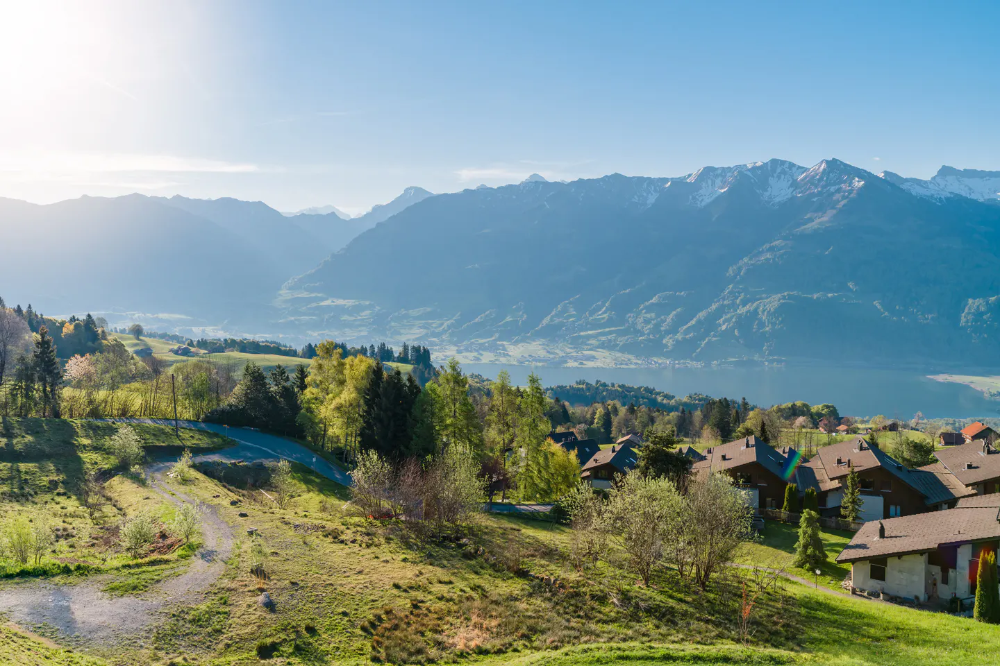 Scenic view of houses on a green hillside, with snow-capped mountains and a lake in the background under a clear blue sky.