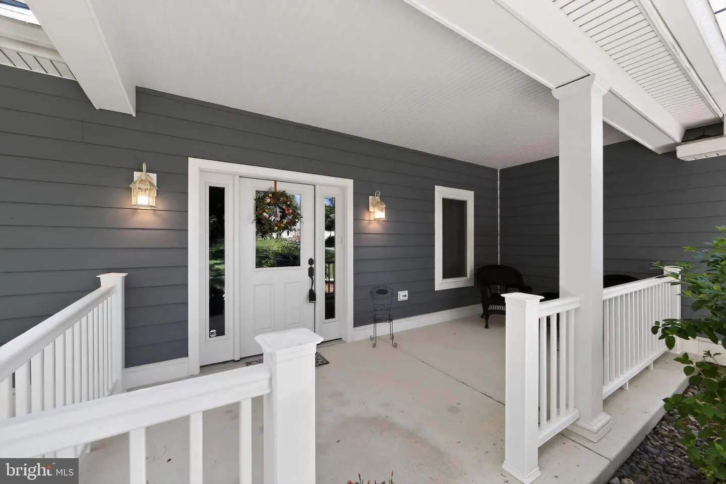 Covered porch with gray siding, white trim, and a white front door with a wreath. Railing and chairs are visible.
