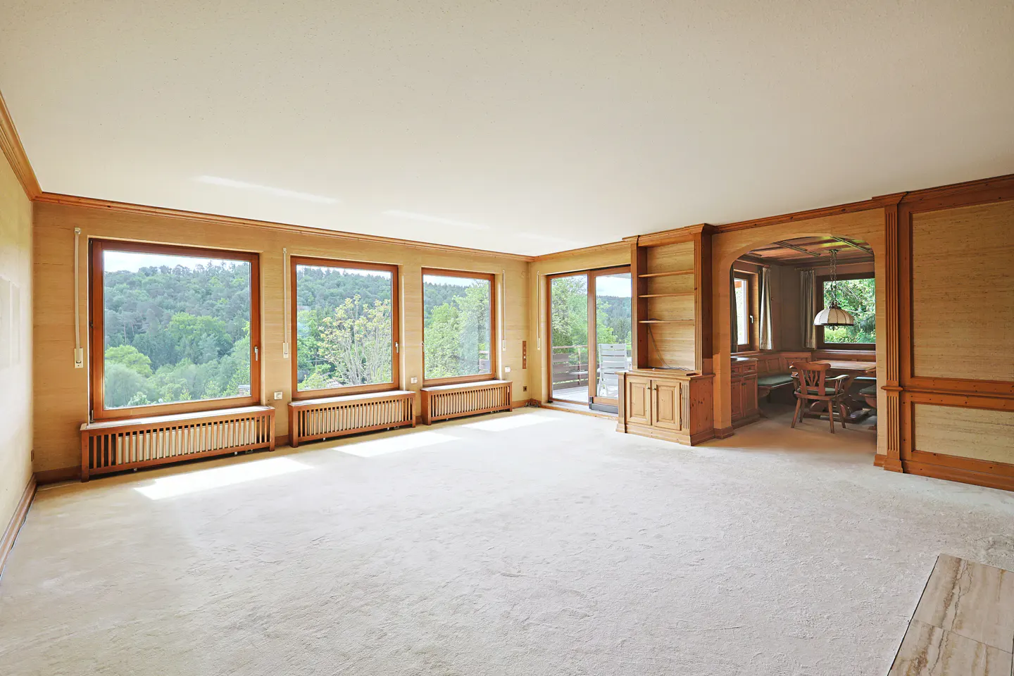 Spacious living room with light carpet, wood-paneled walls, large windows overlooking trees, and an arched dining nook.