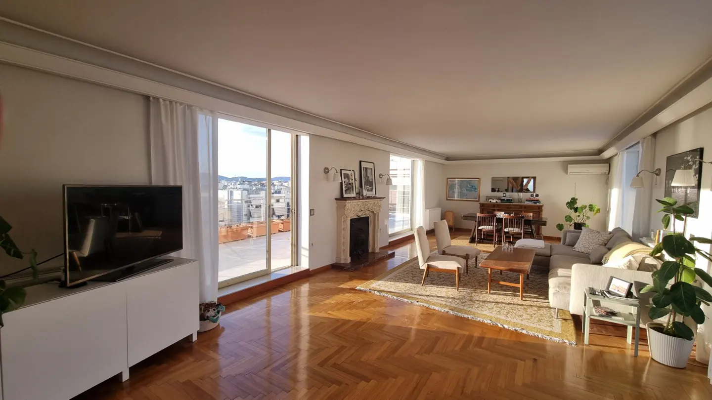 Bright living room with herringbone wood floors, a fireplace, and a large window overlooking a city rooftop. A TV sits on a white cabinet.