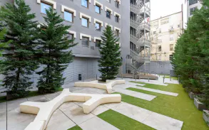 Outdoor courtyard with modern benches, green grass, and evergreen trees. A gray building with windows and a metal fire escape are in the background.