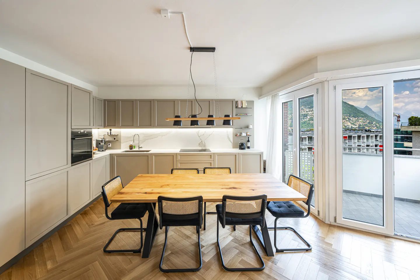 Bright kitchen with wood table, black chairs, and herringbone floor. Gray cabinets line the wall, and a balcony offers a mountain view.