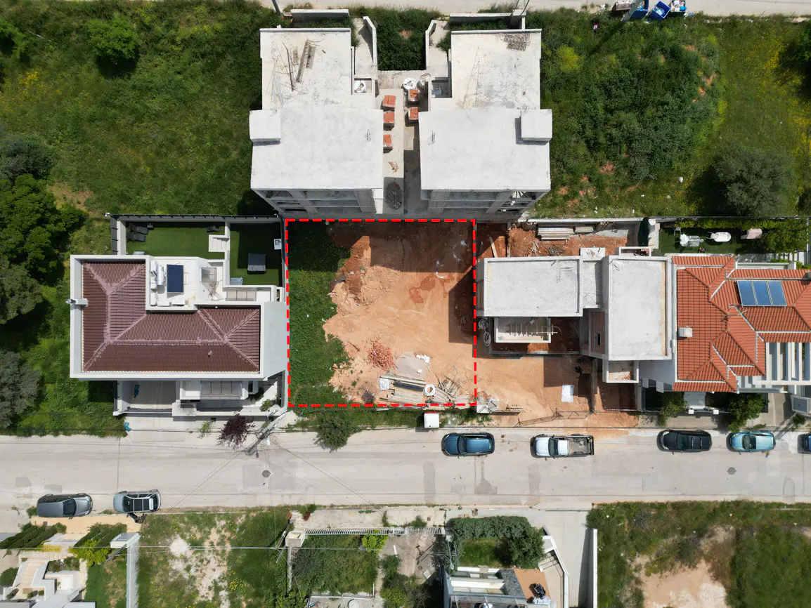 Aerial view of a vacant lot outlined in red, surrounded by houses, a street with parked cars, and green vegetation.