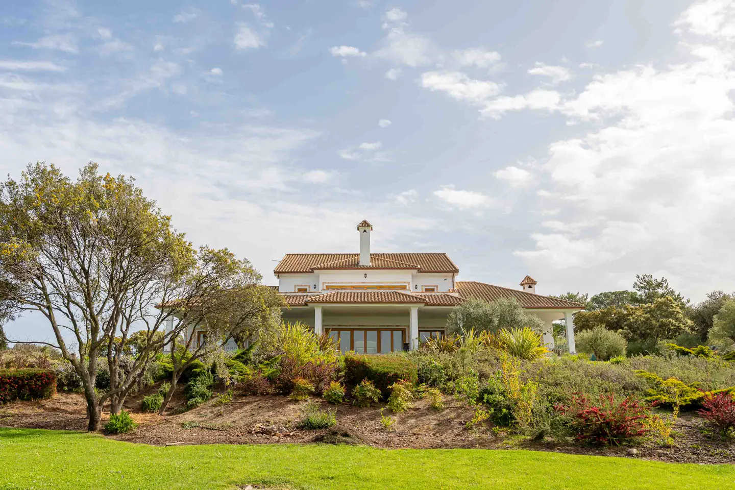 A white house with a red tile roof sits on a hill with green grass and trees.