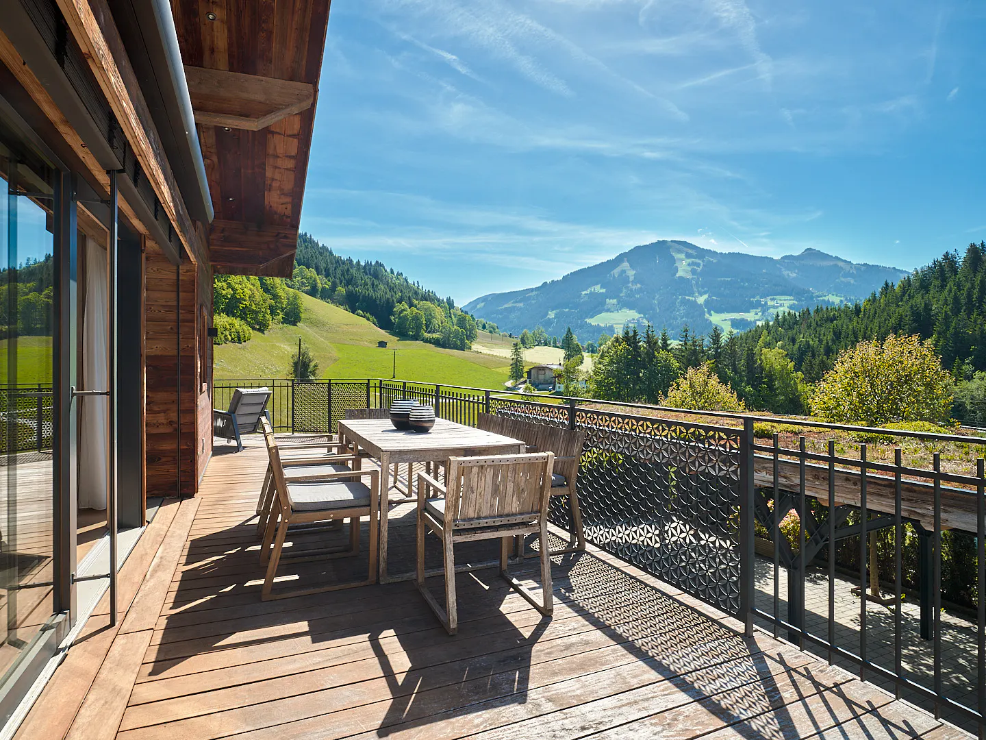 Wooden deck with table and chairs overlooks green hills and mountains under a blue sky. Black metal railing with floral pattern.