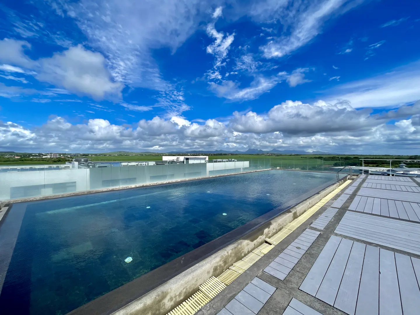 Rooftop pool with blue water reflecting the sky, surrounded by a tiled deck and a glass barrier overlooking a green landscape.