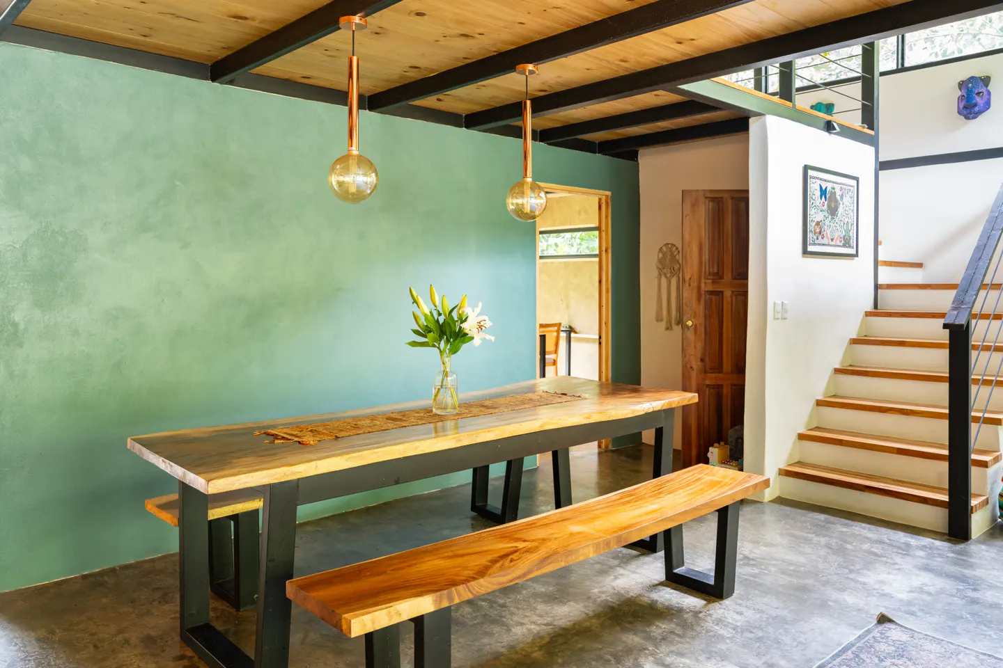 Interior view of a dining area with a wooden table, benches, and a vase of white flowers. A staircase is visible in the background.