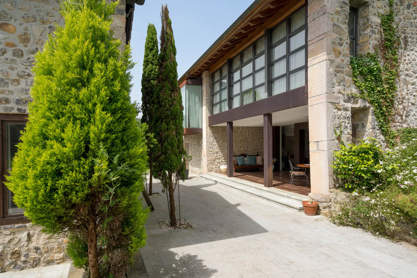 Exterior view of a stone house with large windows, a covered patio, and green trees.
