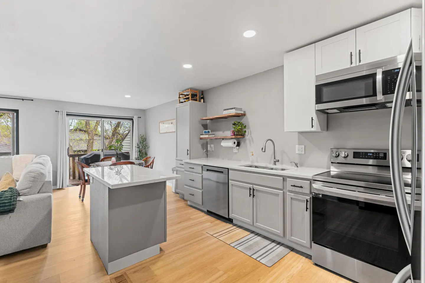 Open concept kitchen and living room with gray cabinets, stainless steel appliances, and light wood floors. A gray sofa is visible in the living room.