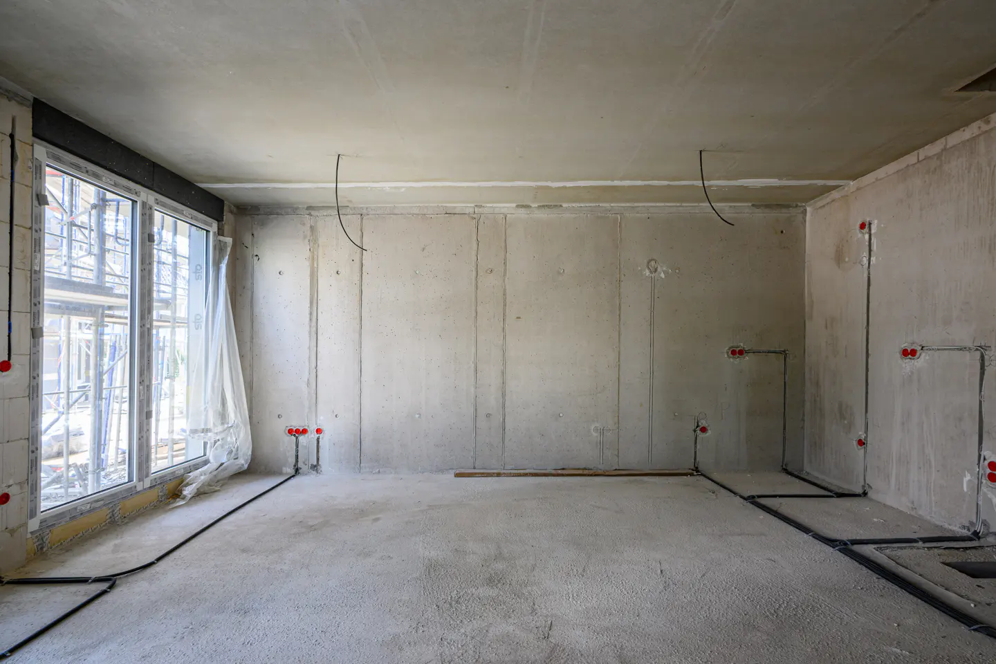 Unfinished room with concrete walls, floor, and ceiling. Large window with scaffolding outside. Electrical wiring and outlets visible.