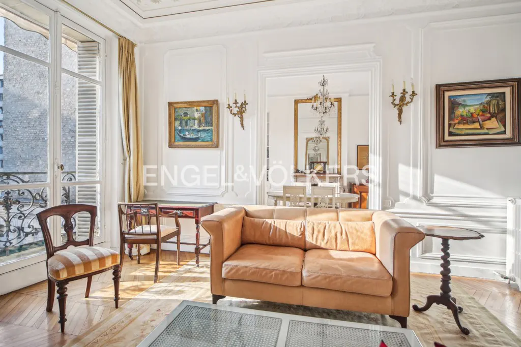 Elegant living room with a tan leather sofa, striped chair, antique desk, and white walls with classic molding.