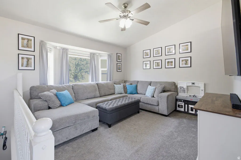 Living room with a large gray sectional, tufted ottoman, blue accent pillows, wall photos, and a ceiling fan.
