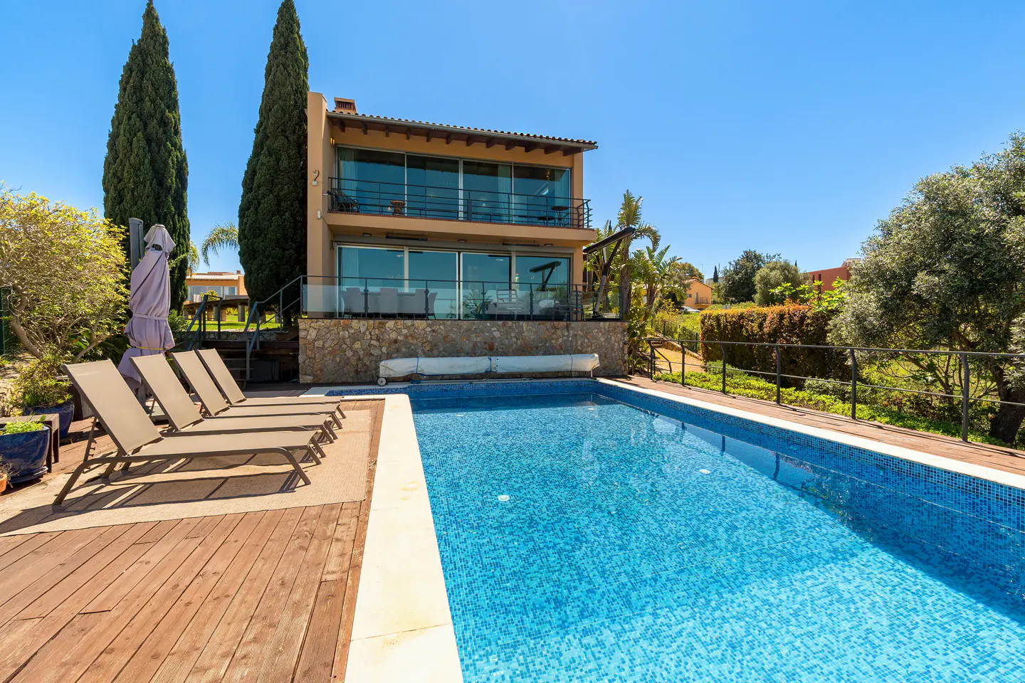 Two-story house with a blue tiled pool, wooden deck, and four lounge chairs. Tall trees and green foliage surround the property.