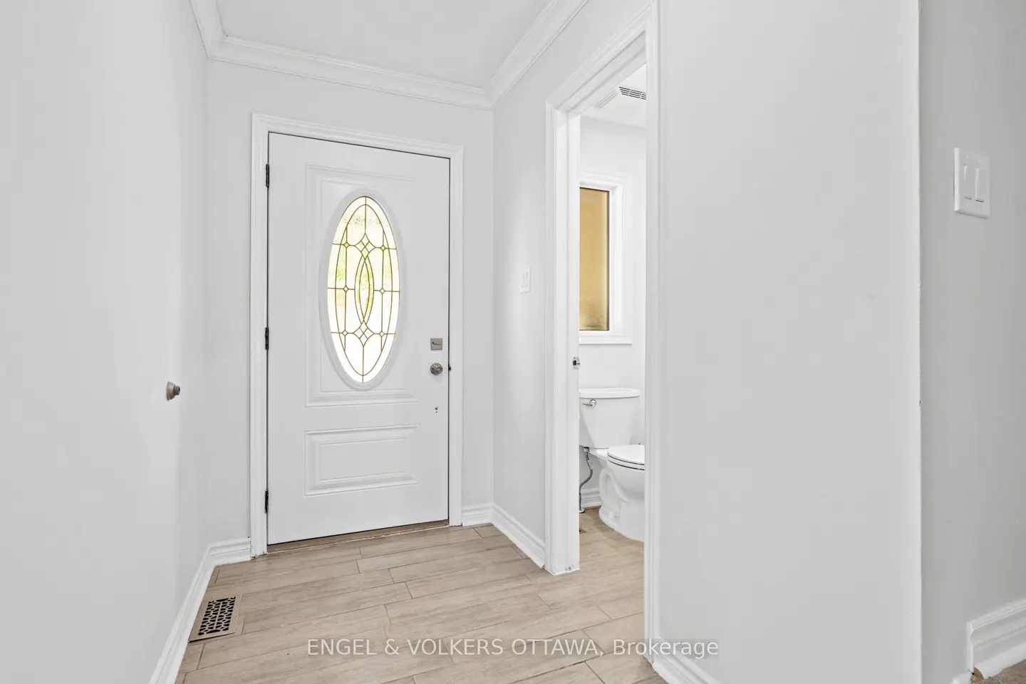 Bright foyer with white walls, light wood-look tile floor, and a white front door with an oval window. A bathroom is visible through an open doorway.