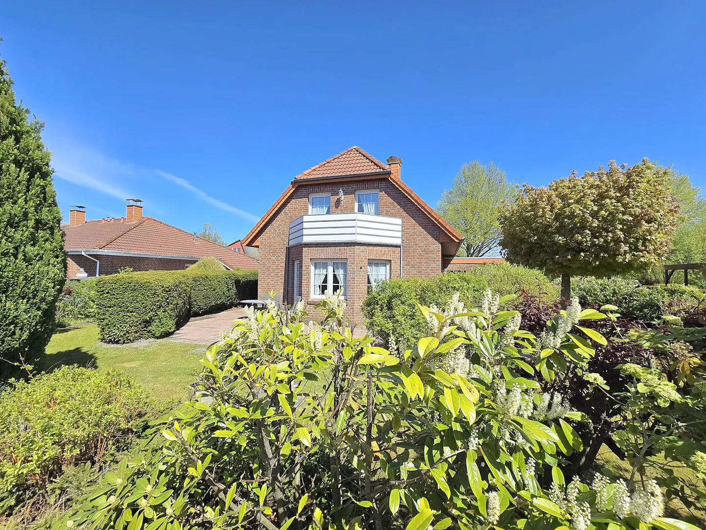 A two-story brick house with a red tile roof and white balcony, surrounded by lush green landscaping under a clear blue sky.
