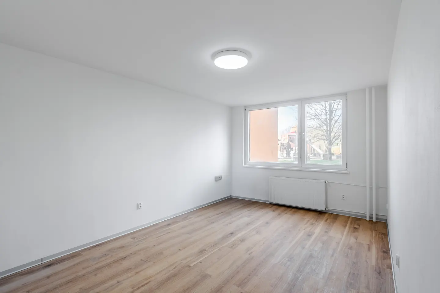 Bright, empty room with wood-look flooring, white walls, and a large window showing a playground outside.