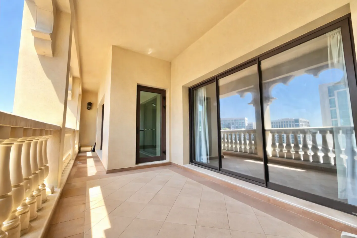 A beige balcony with a tile floor, a decorative balustrade, and a sliding glass door with a view of buildings in the distance.