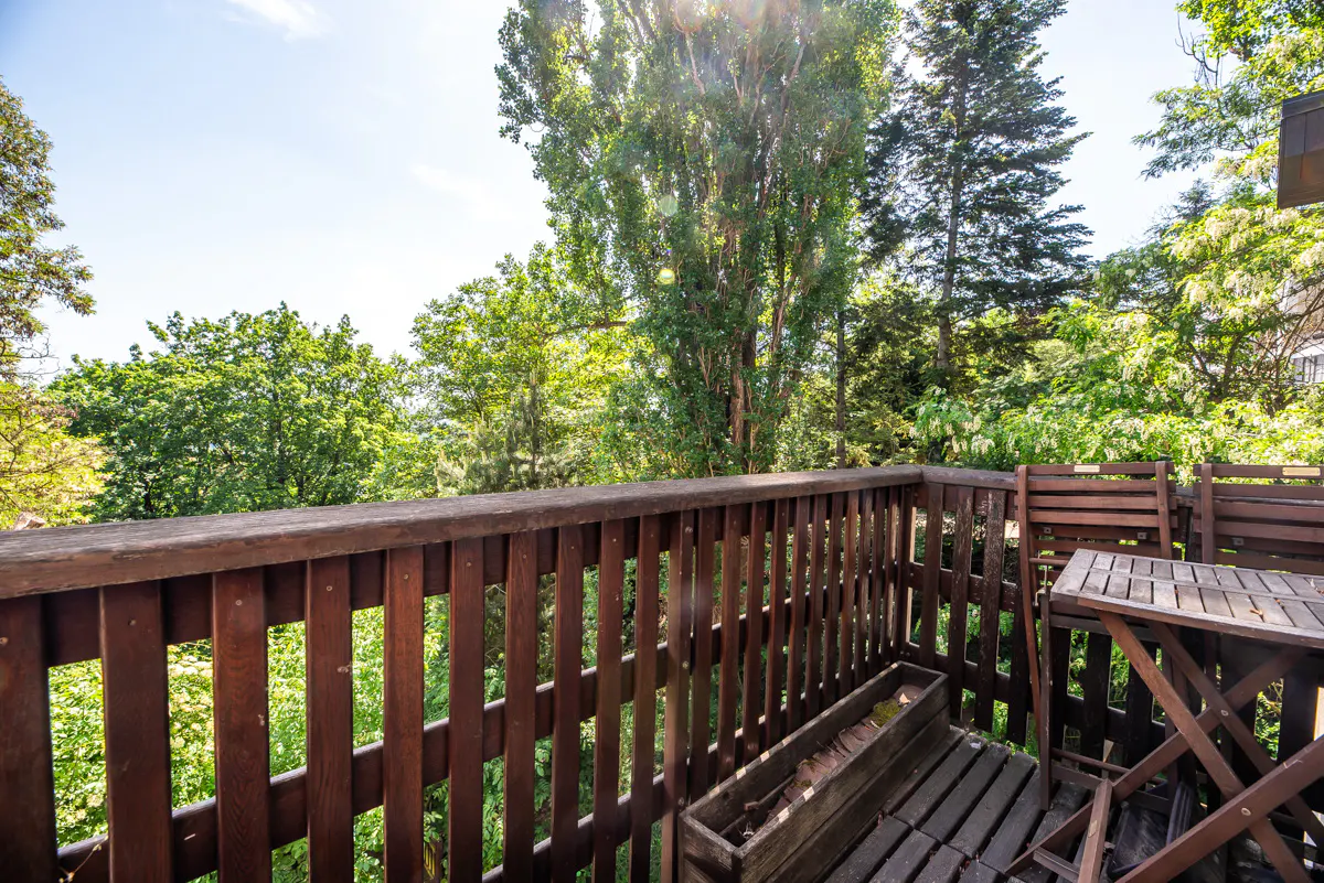 Wooden balcony with a small table and chairs, overlooking a lush green forest under a bright, sunny sky.