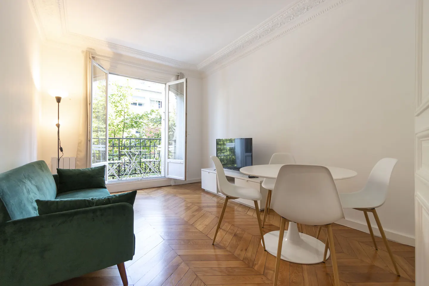 Bright living room with herringbone wood floors, green velvet sofa, white table and chairs, and balcony with open doors.