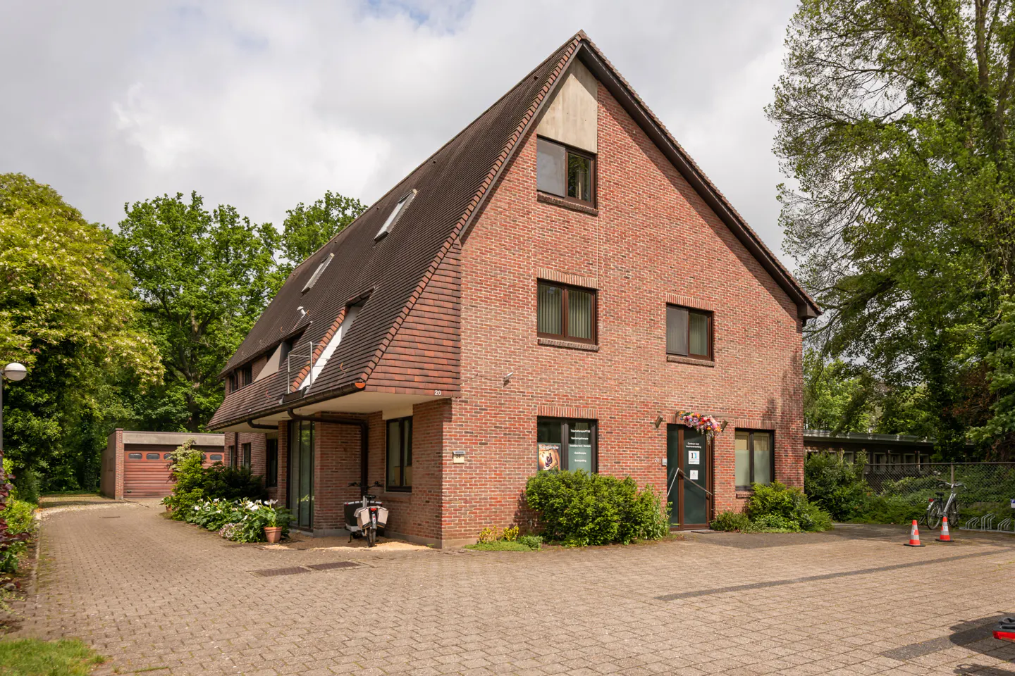 A three-story red brick building with a steep roof, surrounded by trees, with a paved parking area and a small garage.