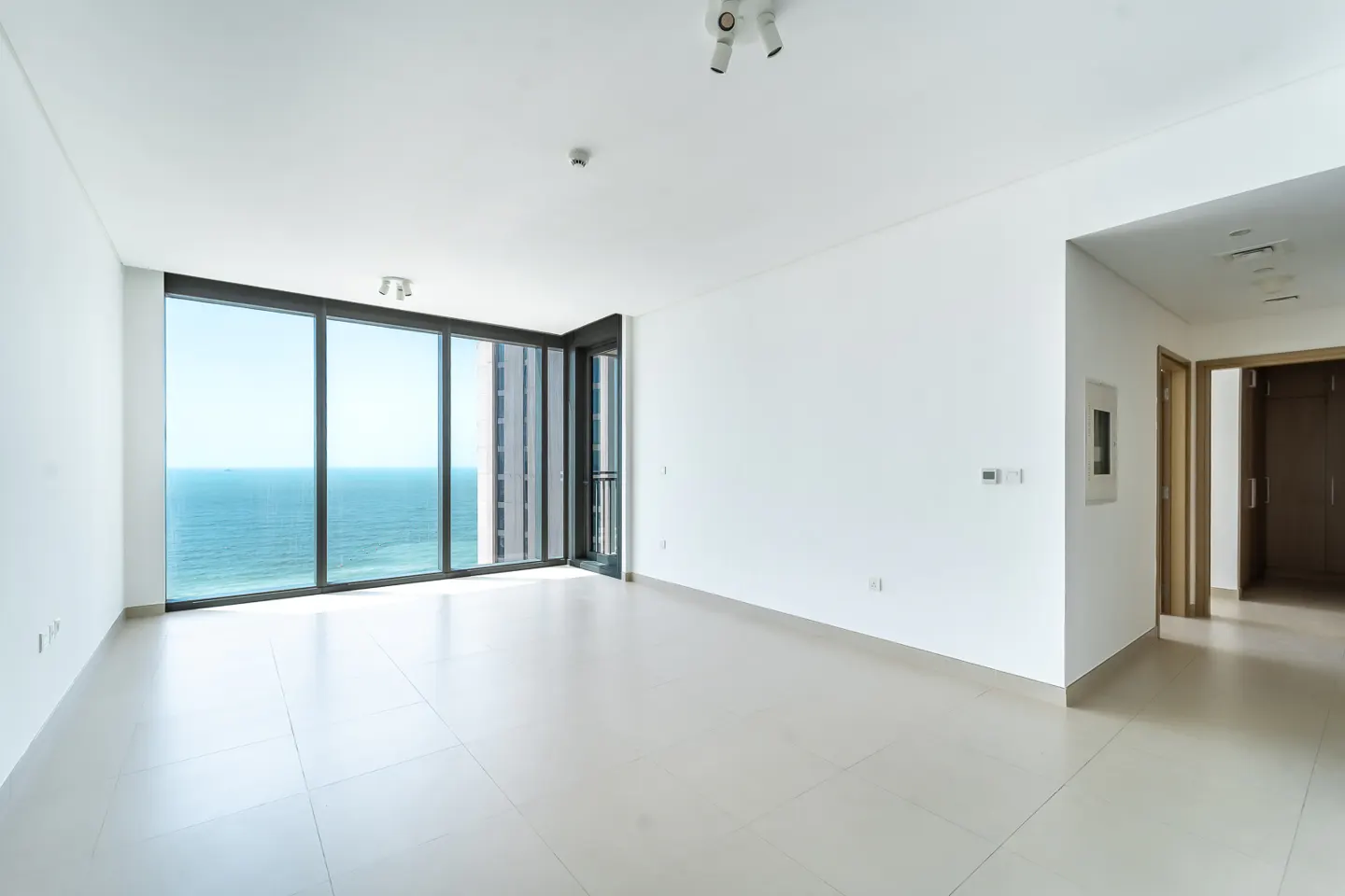 Bright, empty room with white walls and tile floor. Large windows offer ocean views. Hallway with doors visible on the right.