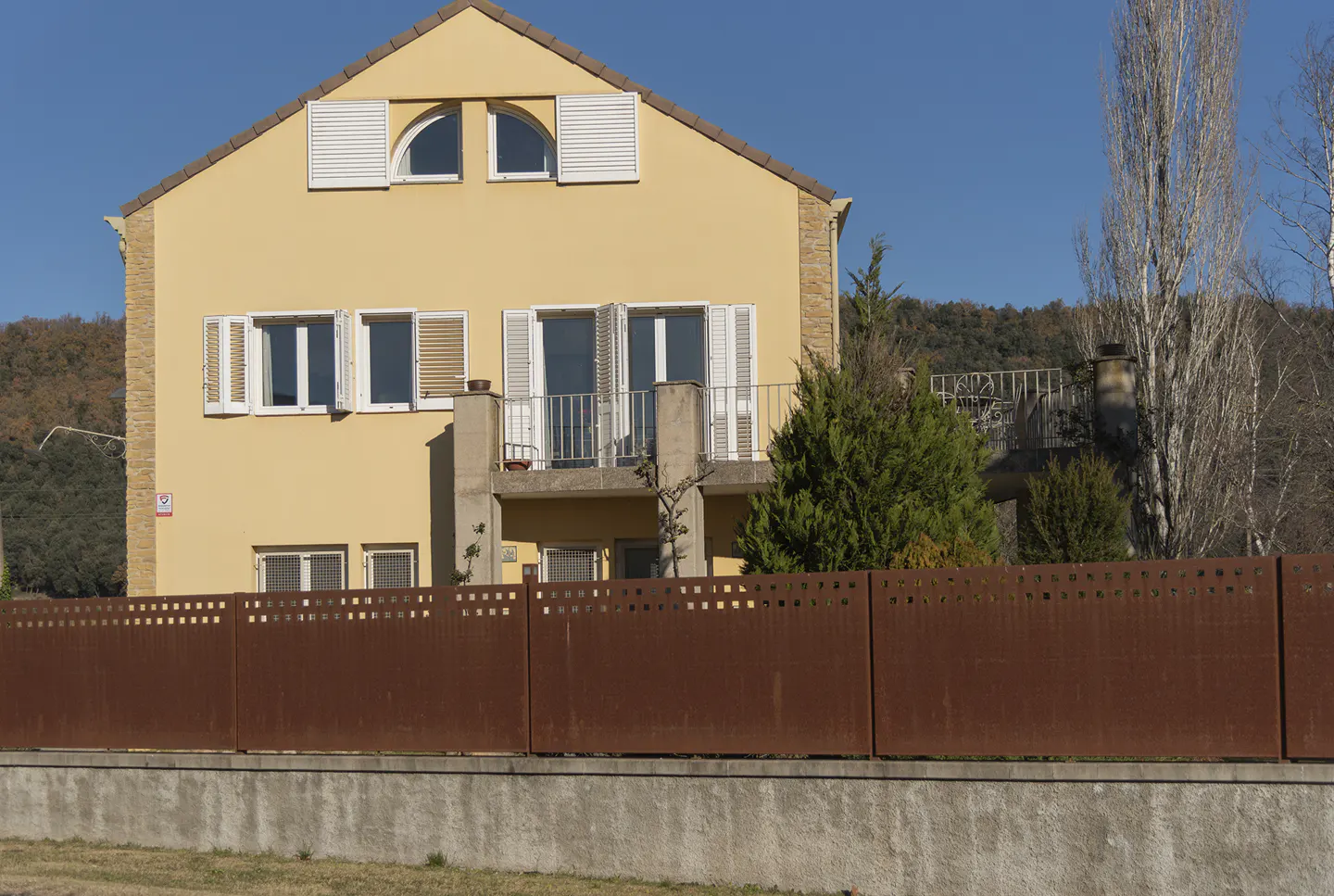 Two-story yellow house with white shutters and a brown metal fence in front. Blue sky.