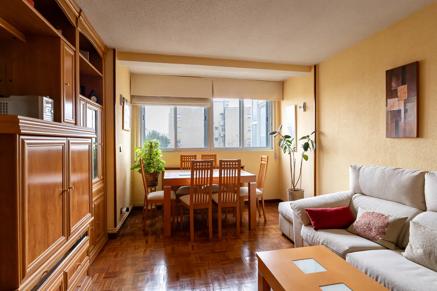 Living room with wood floors, a dining table with six chairs, a large wooden cabinet, and a beige sofa with pillows.