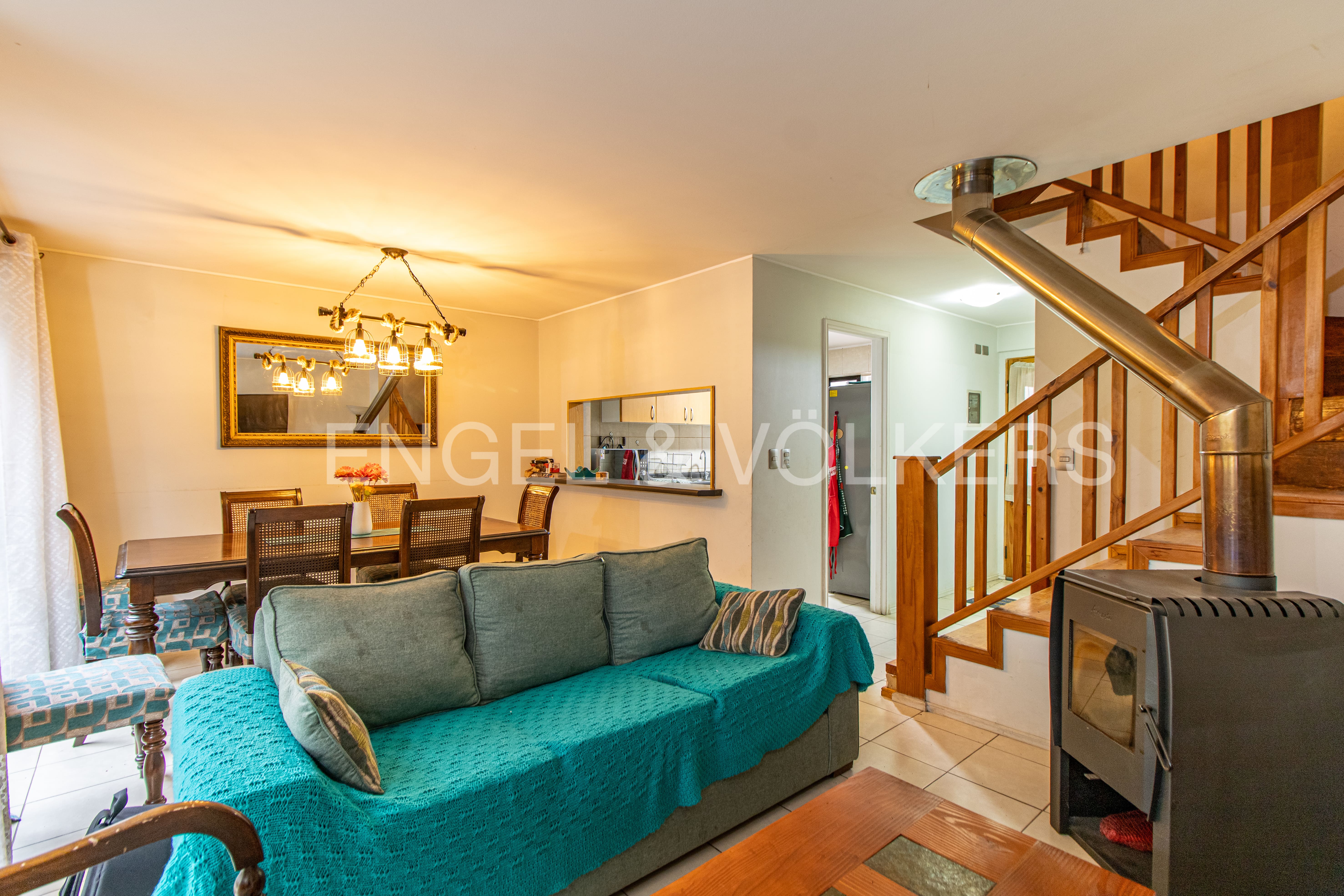 Open-plan living room with a teal sofa, wood-burning stove, wooden staircase, and a dining area with a chandelier.