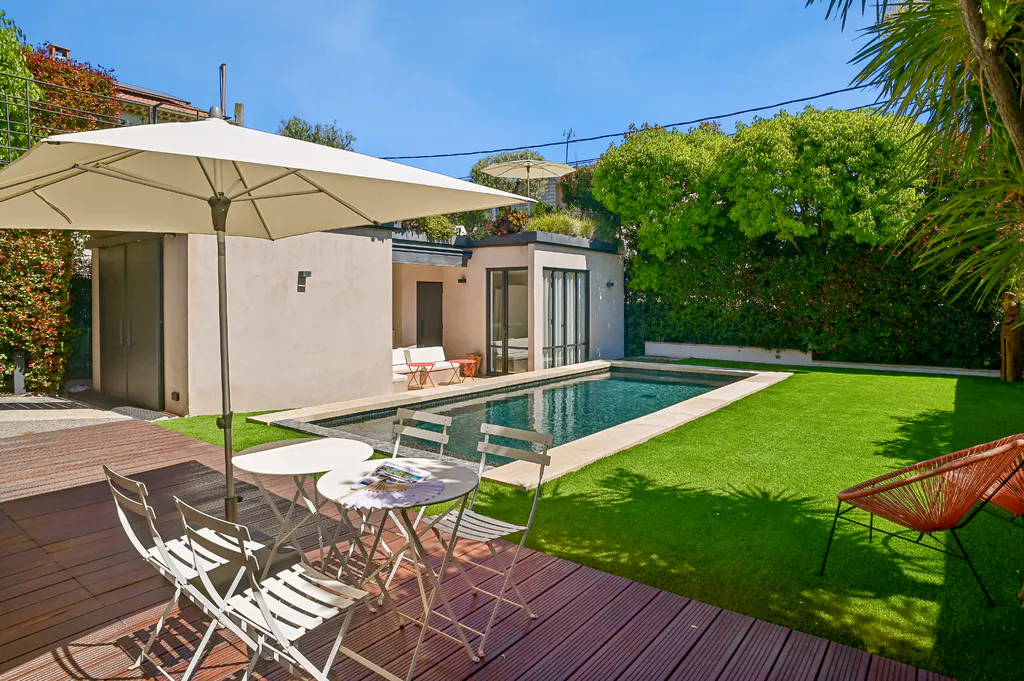 Backyard view with a pool, green lawn, and patio furniture. A white umbrella shades a table and chairs on a wooden deck.