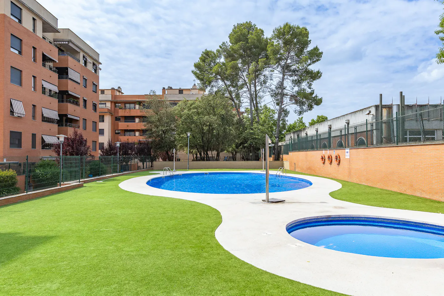Two blue swimming pools surrounded by green artificial grass, set against a backdrop of apartment buildings and trees.
