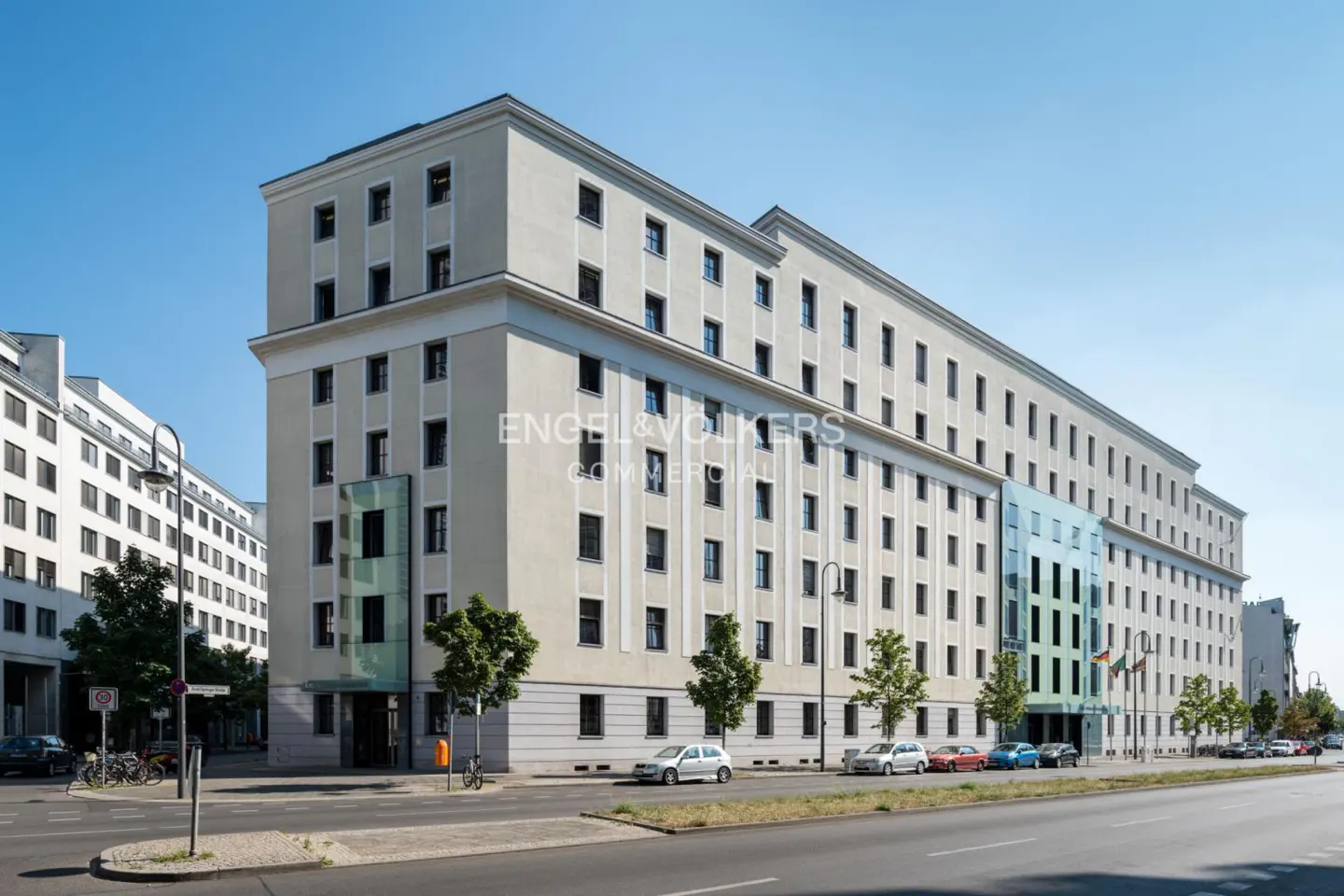 A long, light-beige commercial building with many windows, trees, and cars on a city street.
