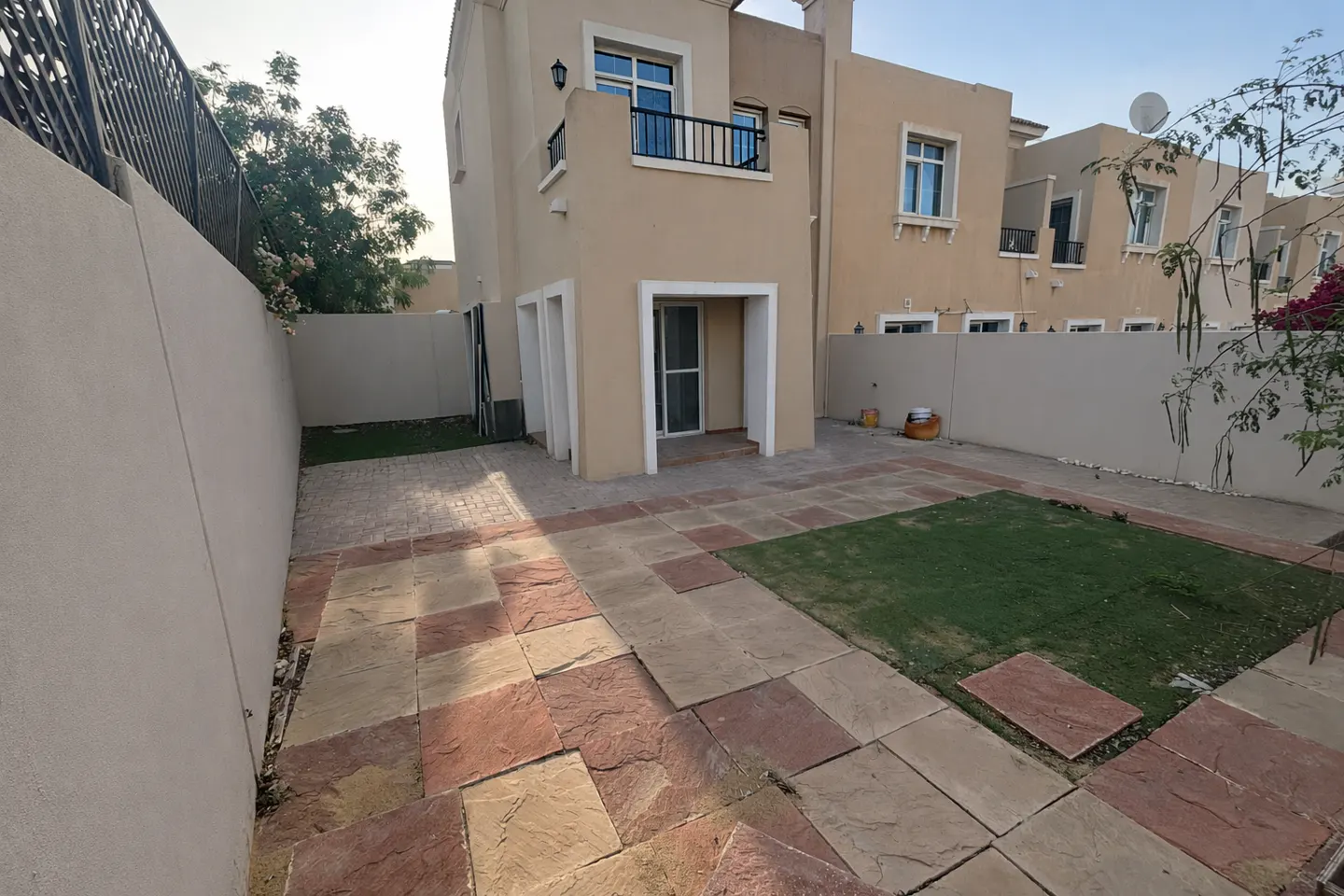 A beige two-story house with a small balcony overlooks a backyard with stone pavers and a patch of green grass.