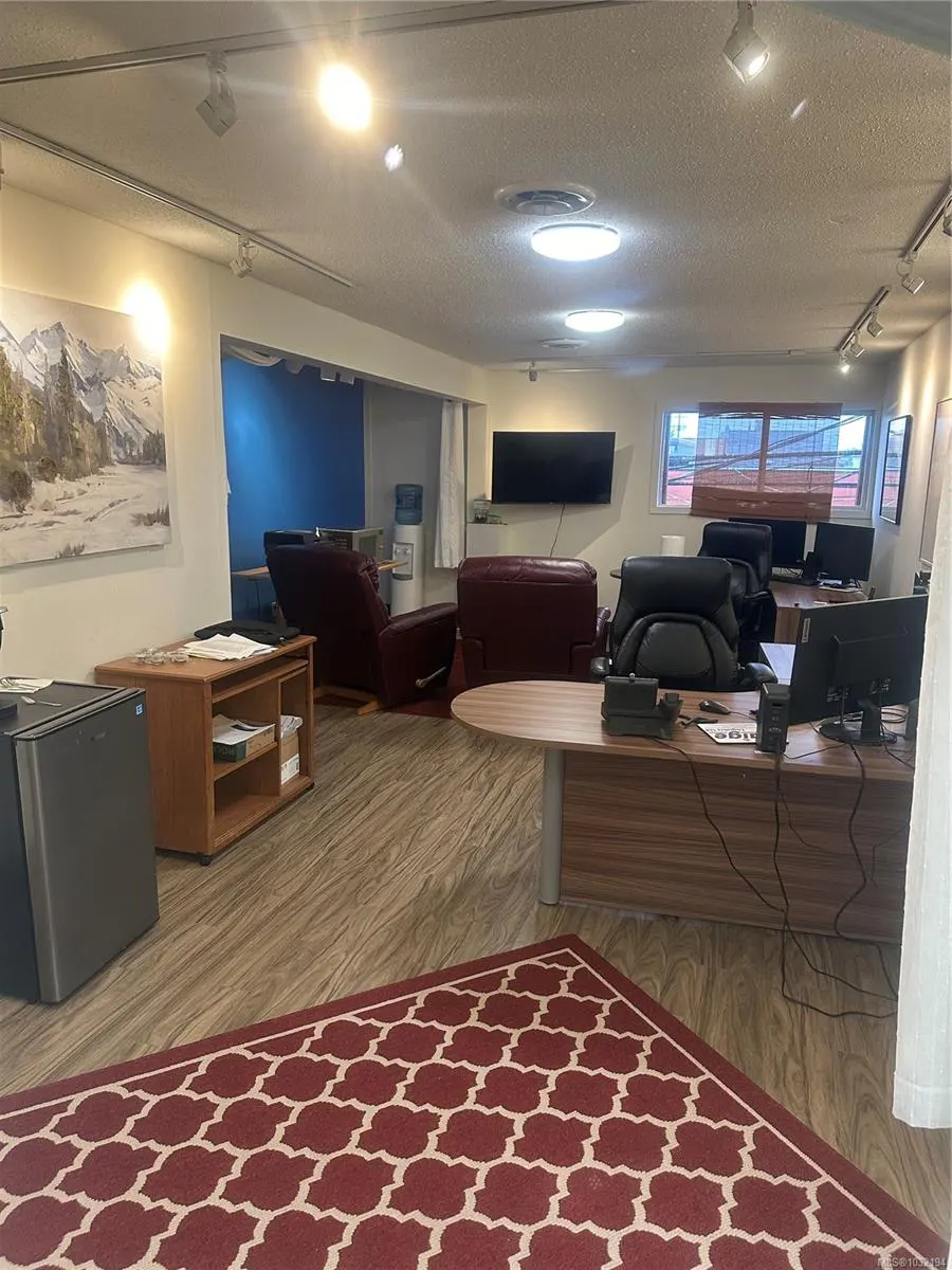 Office space with desks, computers, and chairs. A red patterned rug is on the floor. There are recliners in the background.