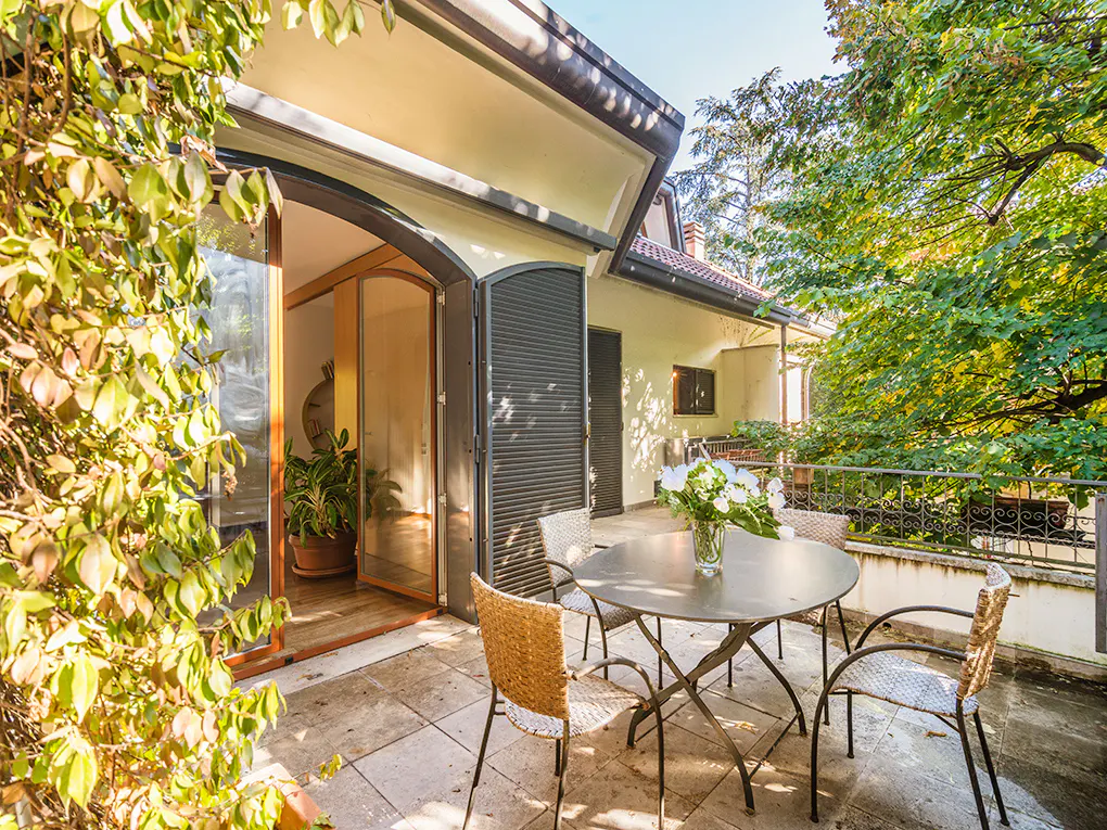 Outdoor patio with a round metal table, four chairs, and a vase of white flowers. Open doors lead inside. Green foliage surrounds the area.