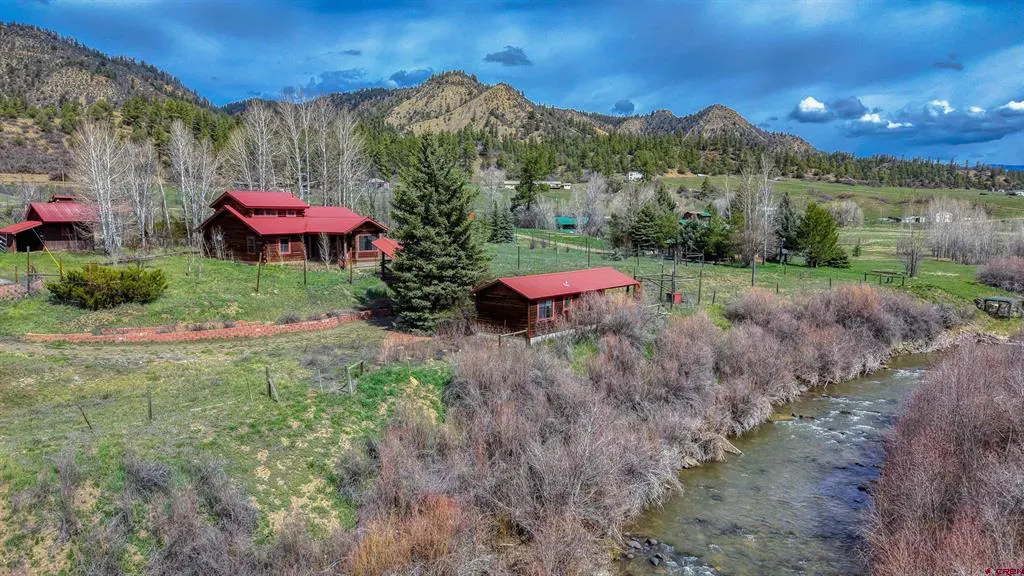 A landscape view of a log cabin with a red roof next to a river and mountains.
