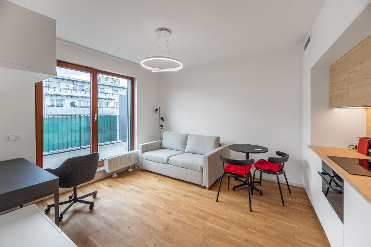 Modern studio apartment with light wood floors, a grey sofa, a small dining table with red cushions, and a balcony view.