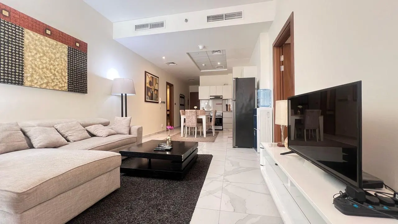 A bright apartment living room with a beige sofa, black coffee table, and a TV on a white stand. The kitchen and dining area are visible in the background.
