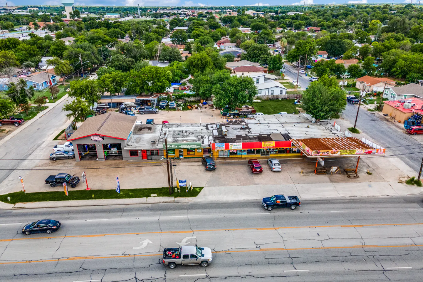 Aerial view of a commercial property with a gray auto shop, colorful storefronts, and a gas station canopy along a cracked road with passing cars.